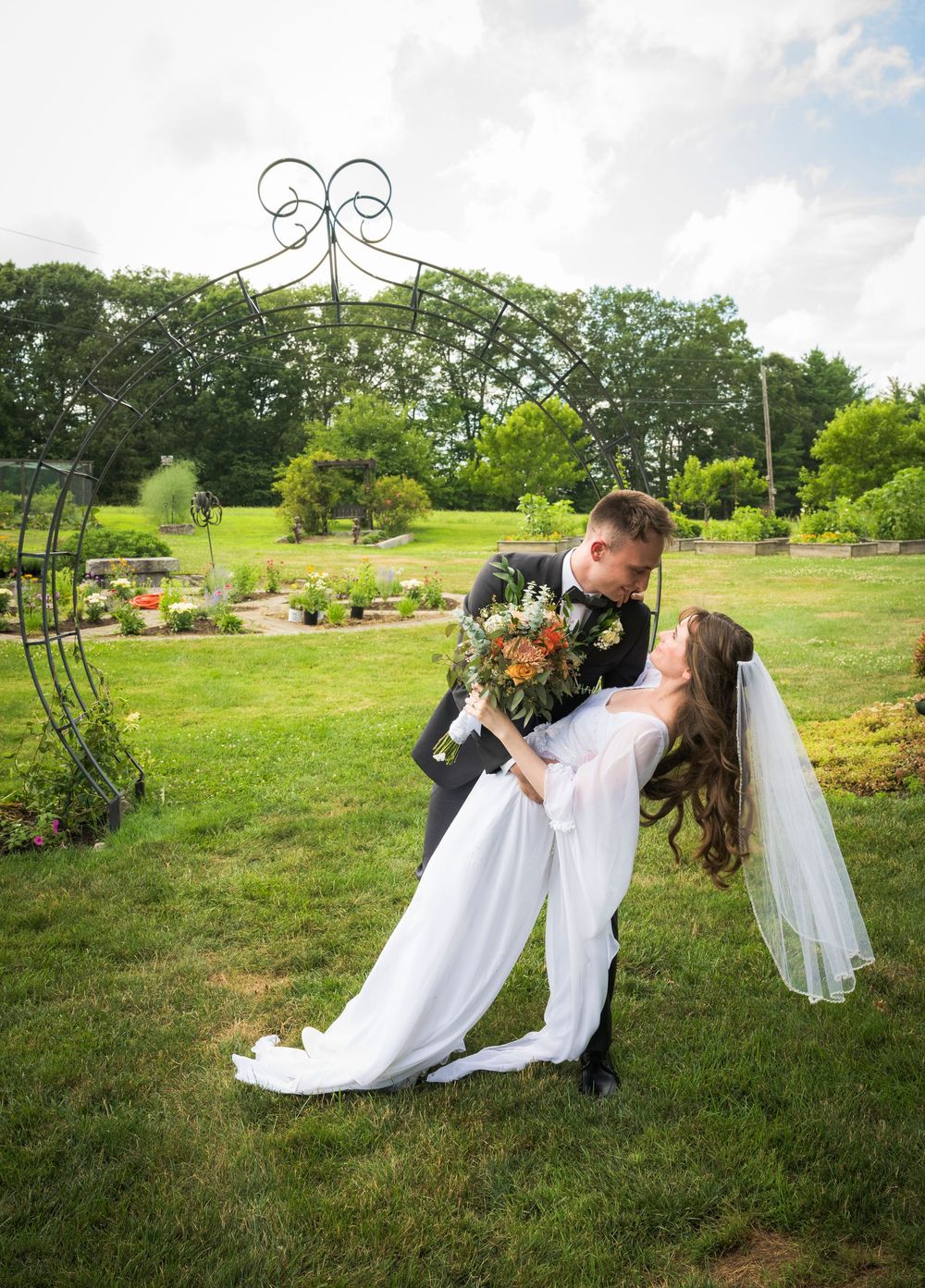 Romantic wedding dip pose under decorative garden arch in lush green landscape.
