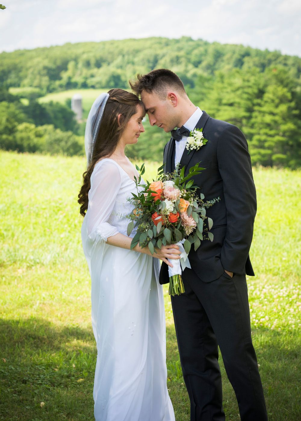 Wedding couple stands in a sunny meadow with wildflowers, surrounded by lush greenery in a romantic outdoor setting.