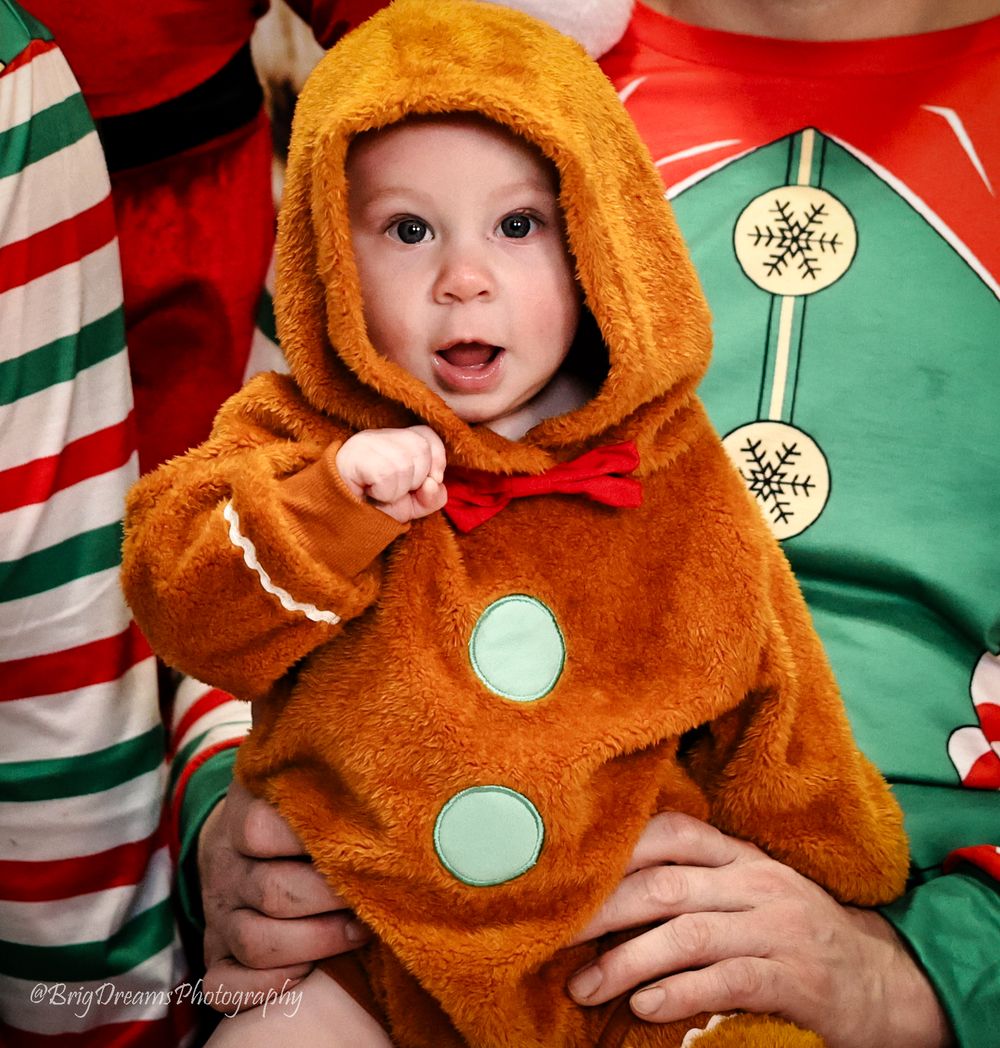 A small child wearing a plush brown gingerbread costume with white buttons sits among holiday-themed clothing.