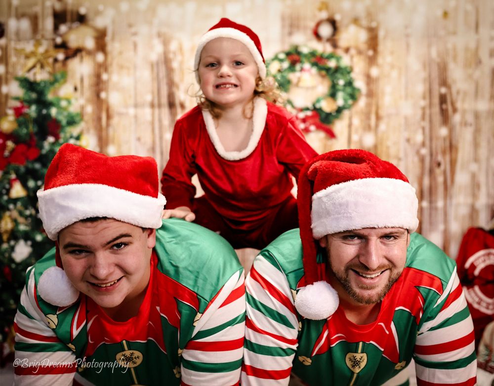 Family poses in matching festive red and green Christmas outfits with Santa hats and striped elf socks against rustic backdrop.