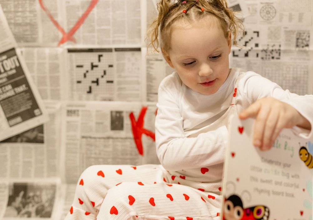 Child in heart-patterned white outfit sits against newspaper wall with red decorative hearts.