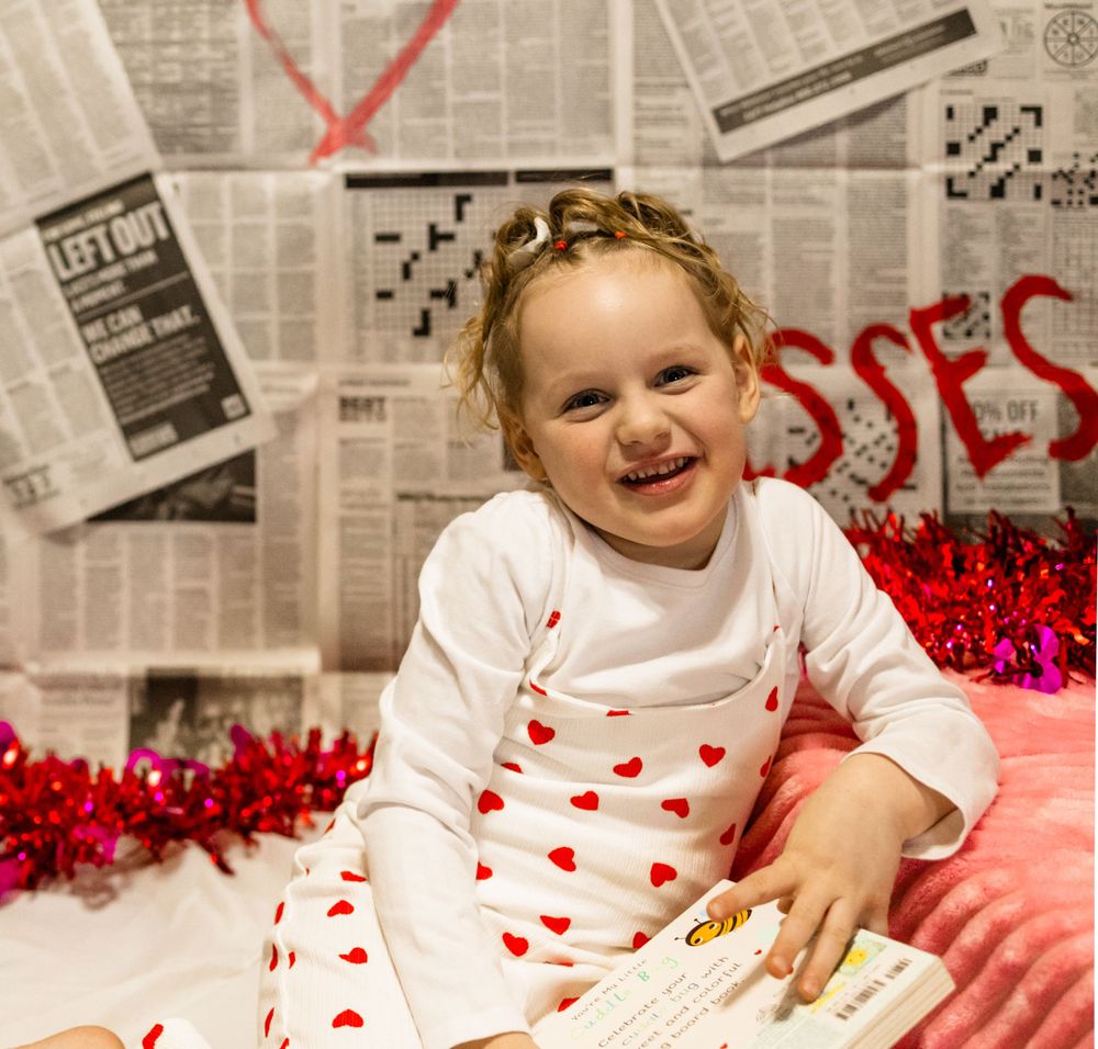 Child in heart-patterned outfit smiles while reading against newspaper-covered wall with red decorations.