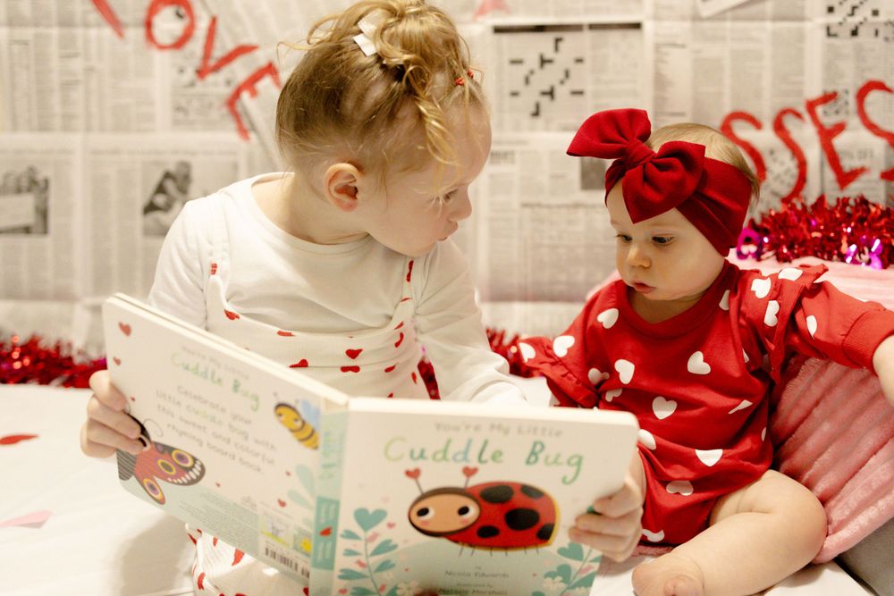 Two children in red and white heart-patterned outfits share a ladybug storybook moment.