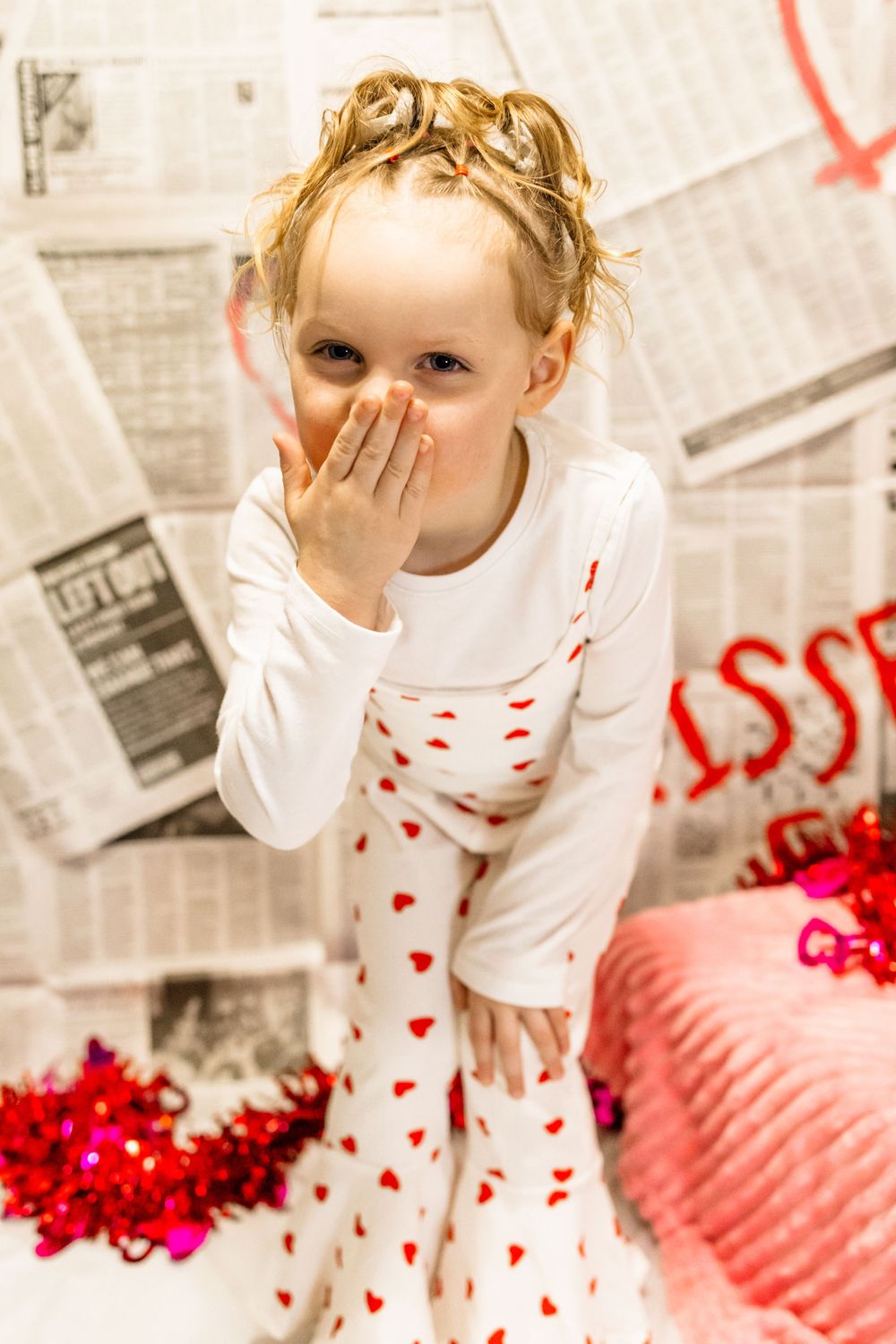 Valentine's themed photo with white heart-print pajamas against newspaper backdrop and red decor.
