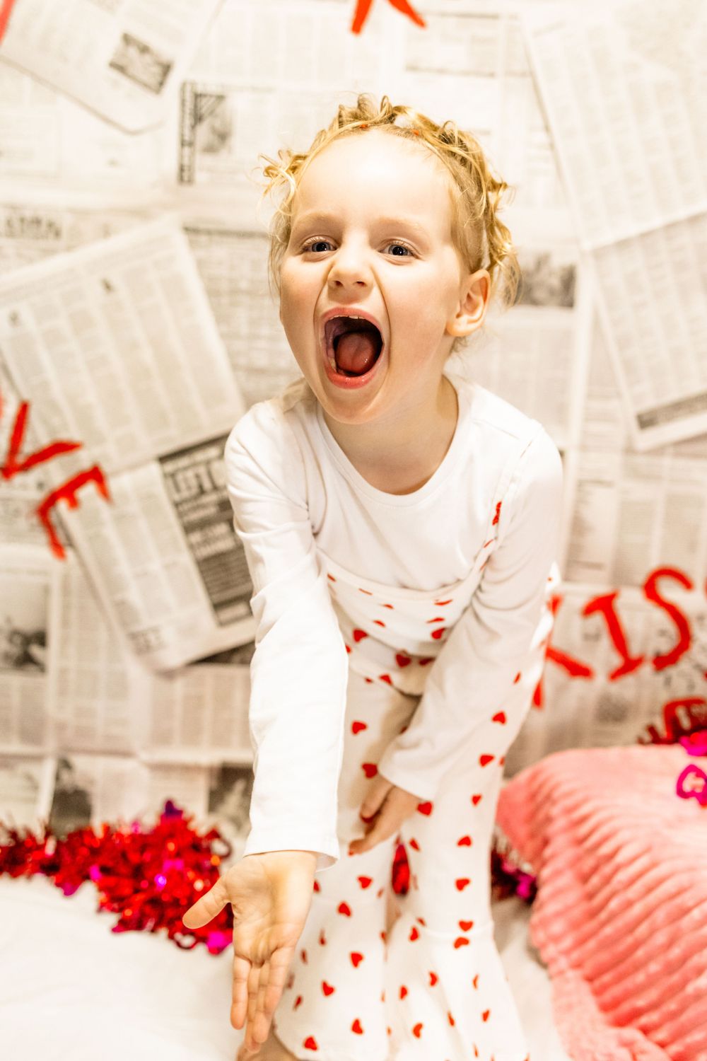 Playful pose in white heart-print pajamas against newspaper backdrop with red decorations.