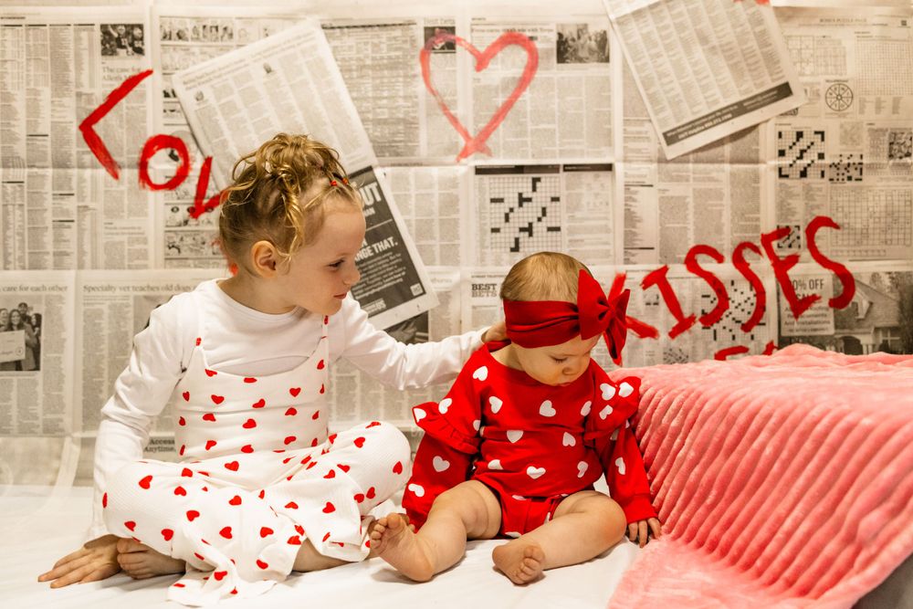 Two children in Valentine's themed outfits sit on white bedding with newspaper backdrop and heart decorations.