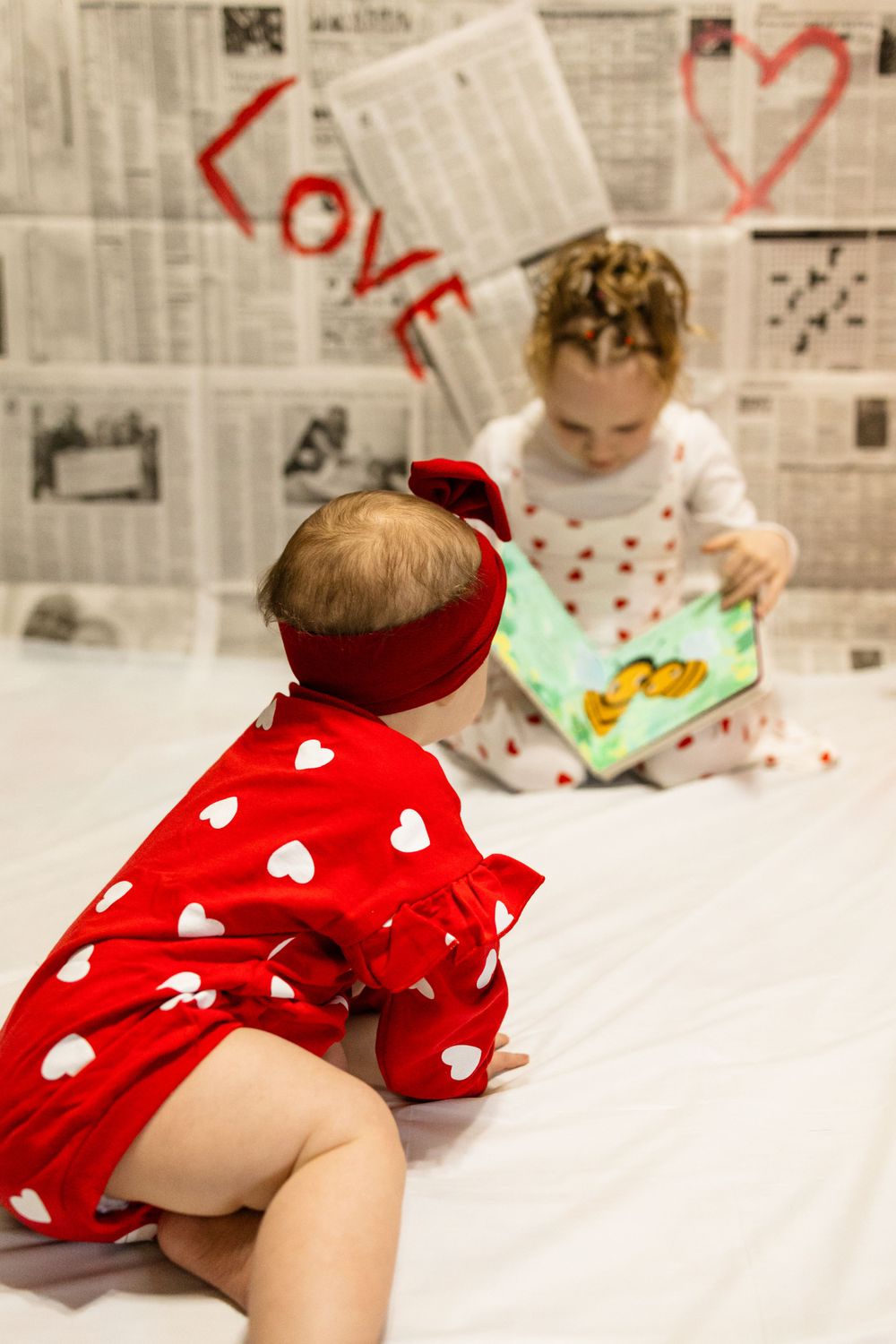 Two children in heart-patterned outfits share a storybook moment against newspaper-covered walls.