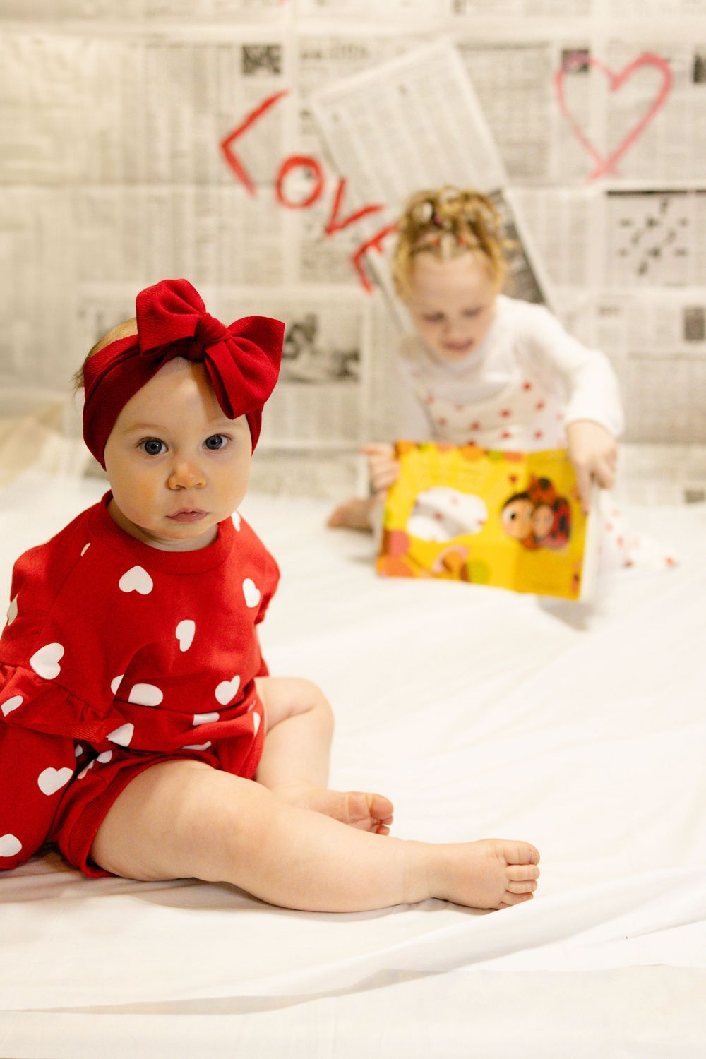Two children in Valentine's themed outfits reading together on white bedding with newspaper wall backdrop.