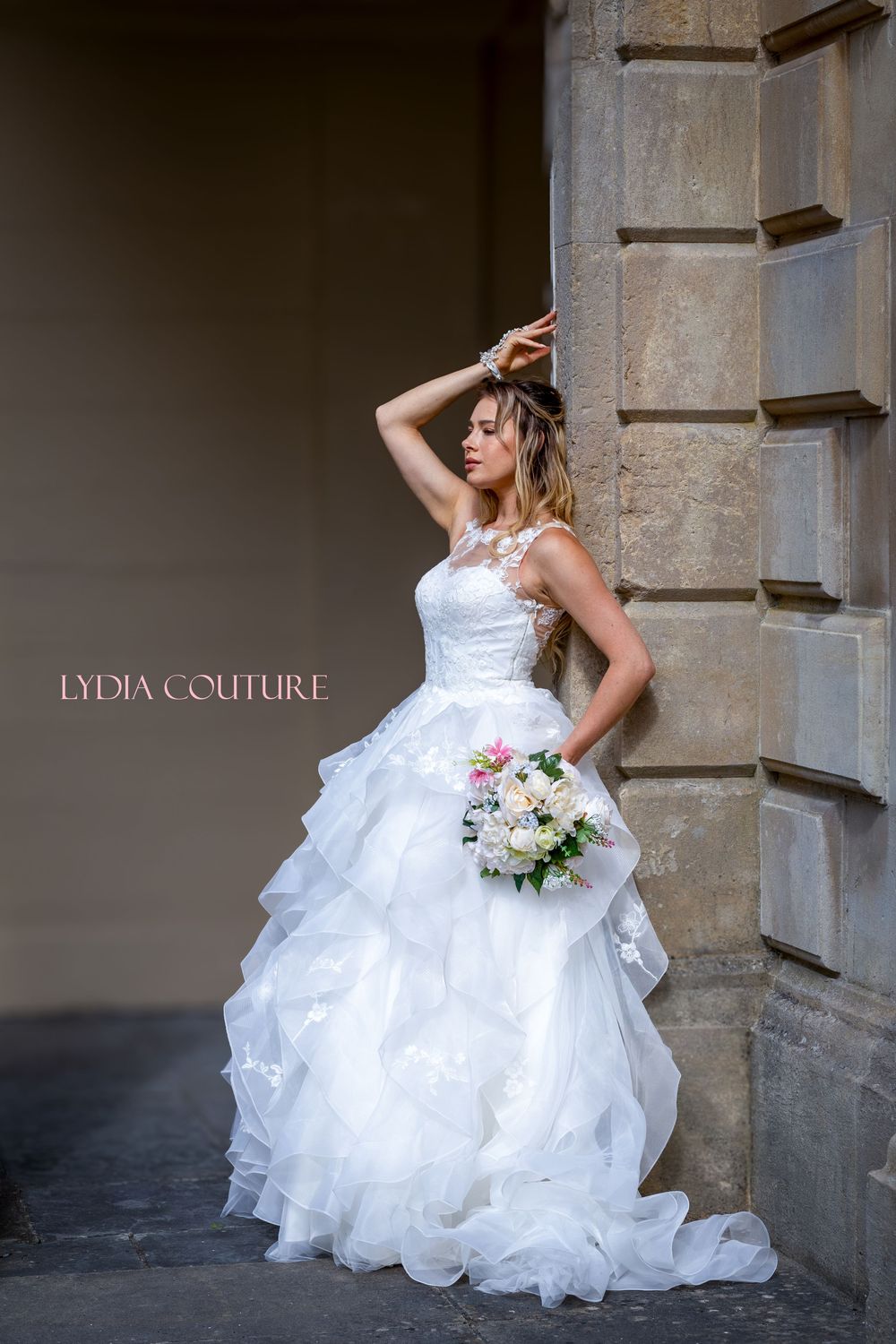 A wedding dress with a layered tulle skirt and lace bodice is showcased against a stone architectural backdrop with floral bouquet.