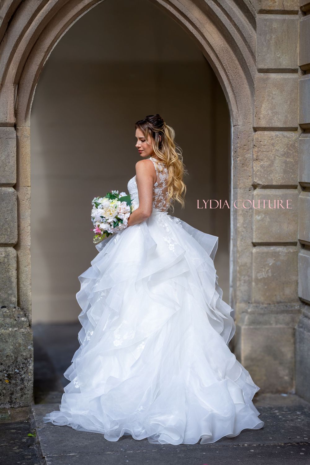 A bride in a luxurious white ruffled wedding gown stands in a stone archway holding a pastel floral bouquet. By Bristol wedding photographer