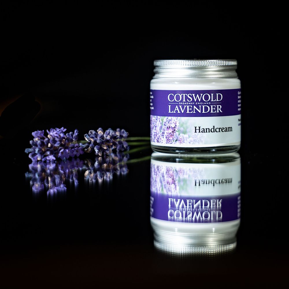 Purple lavender flowers arranged with silver jar of Cotswold handcream against black background.