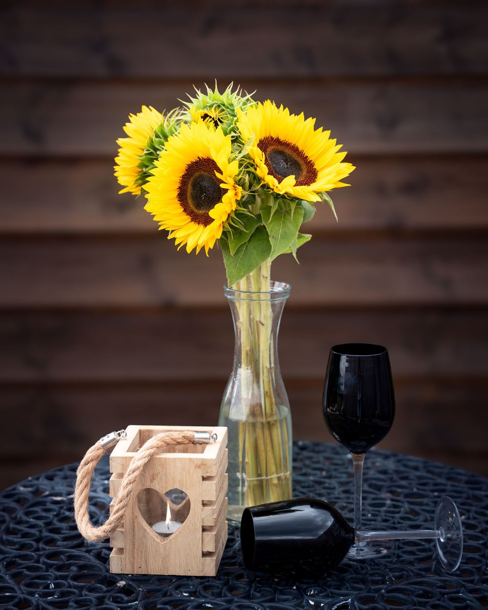 Three vibrant sunflowers in a clear glass vase alongside a black wine glass and wooden candle holder on a dark table.