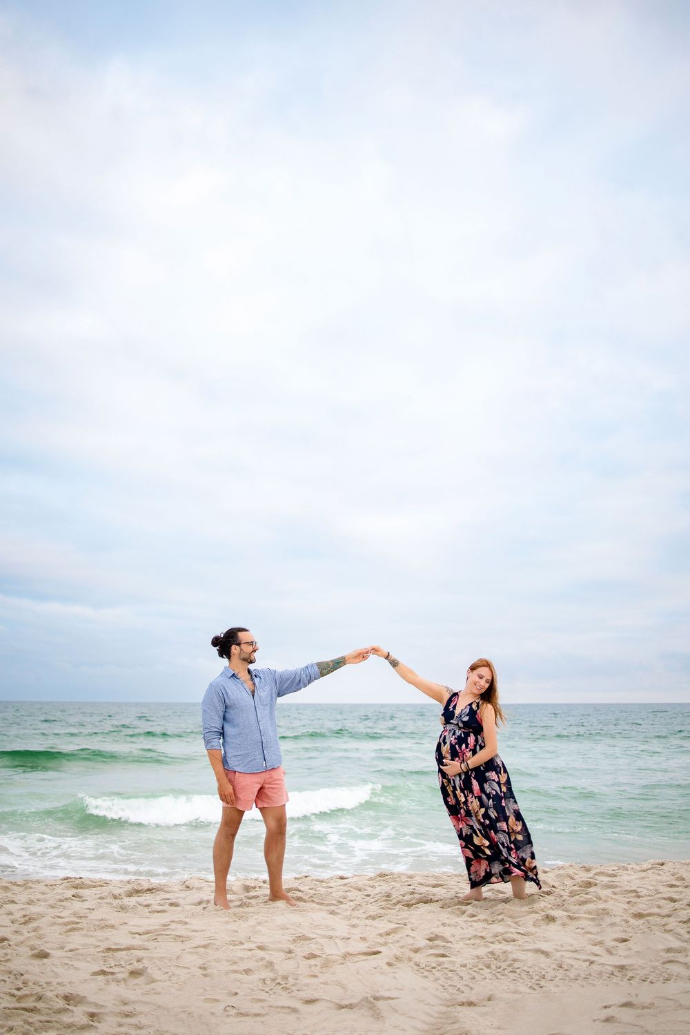 A couple shares a romantic dance pose on the beach with turquoise ocean waves and cloudy sky in the background.