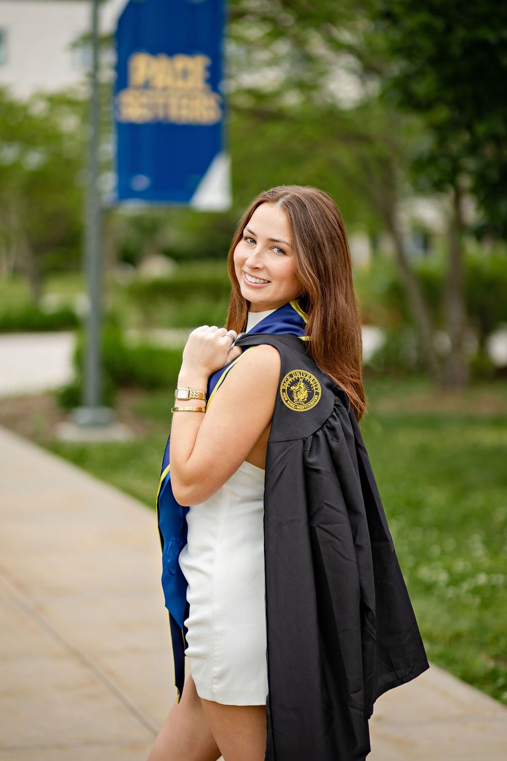 A smiling young woman in a white dress holds her black graduation gown over her shoulder on a college campus.