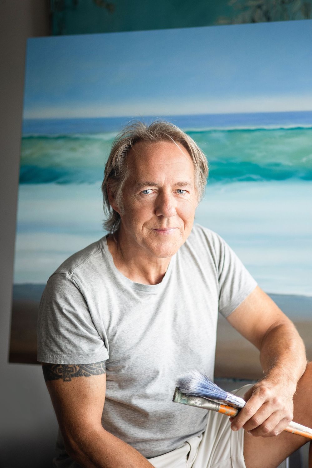 Artist sits in front of coastal painting wearing grey t-shirt in studio setting.