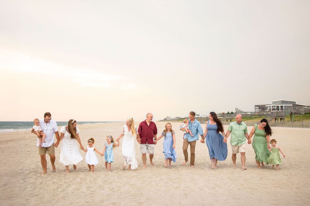 A group in coordinated pastel outfits walks together along a sandy beach at sunset with coastal homes in the background.