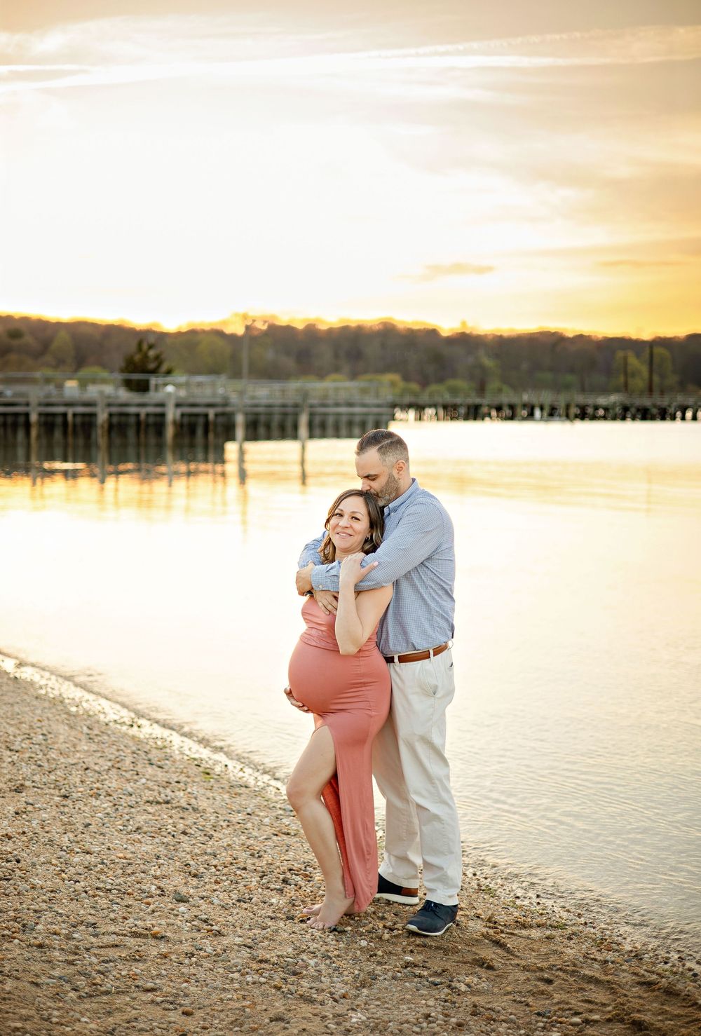 Couple embracing on sandy beach at golden sunset, pregnant woman in pink dress, man in blue shirt, waterfront backdrop.