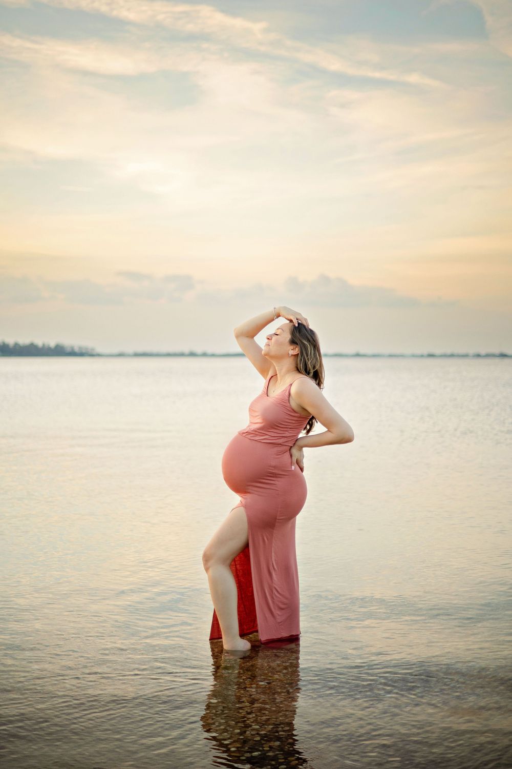 Pregnant woman in pink dress standing in calm water at sunset, posing gracefully with hand on hip.