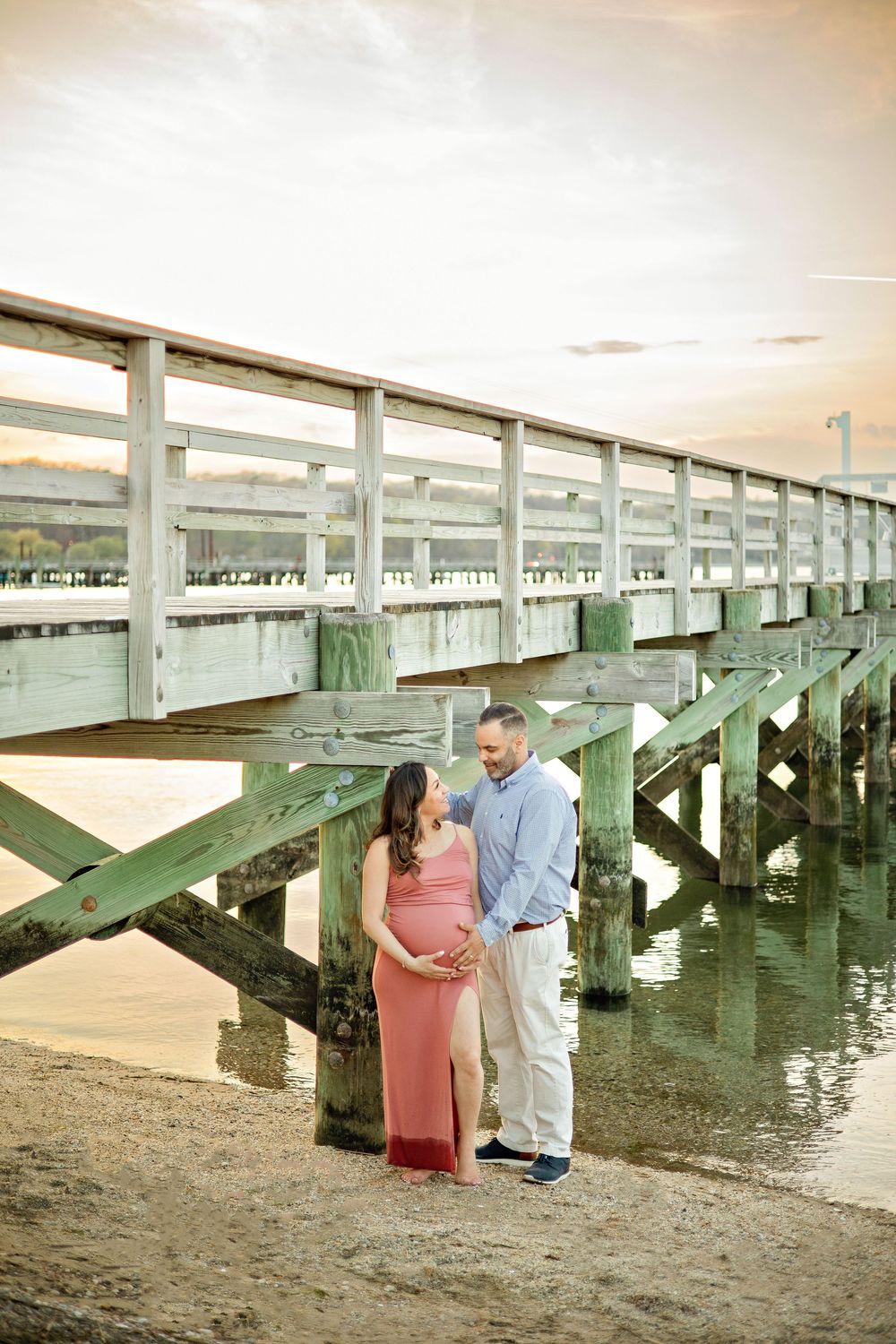 Pregnant couple standing under a wooden pier on a sandy beach at sunset, woman in pink maternity dress.