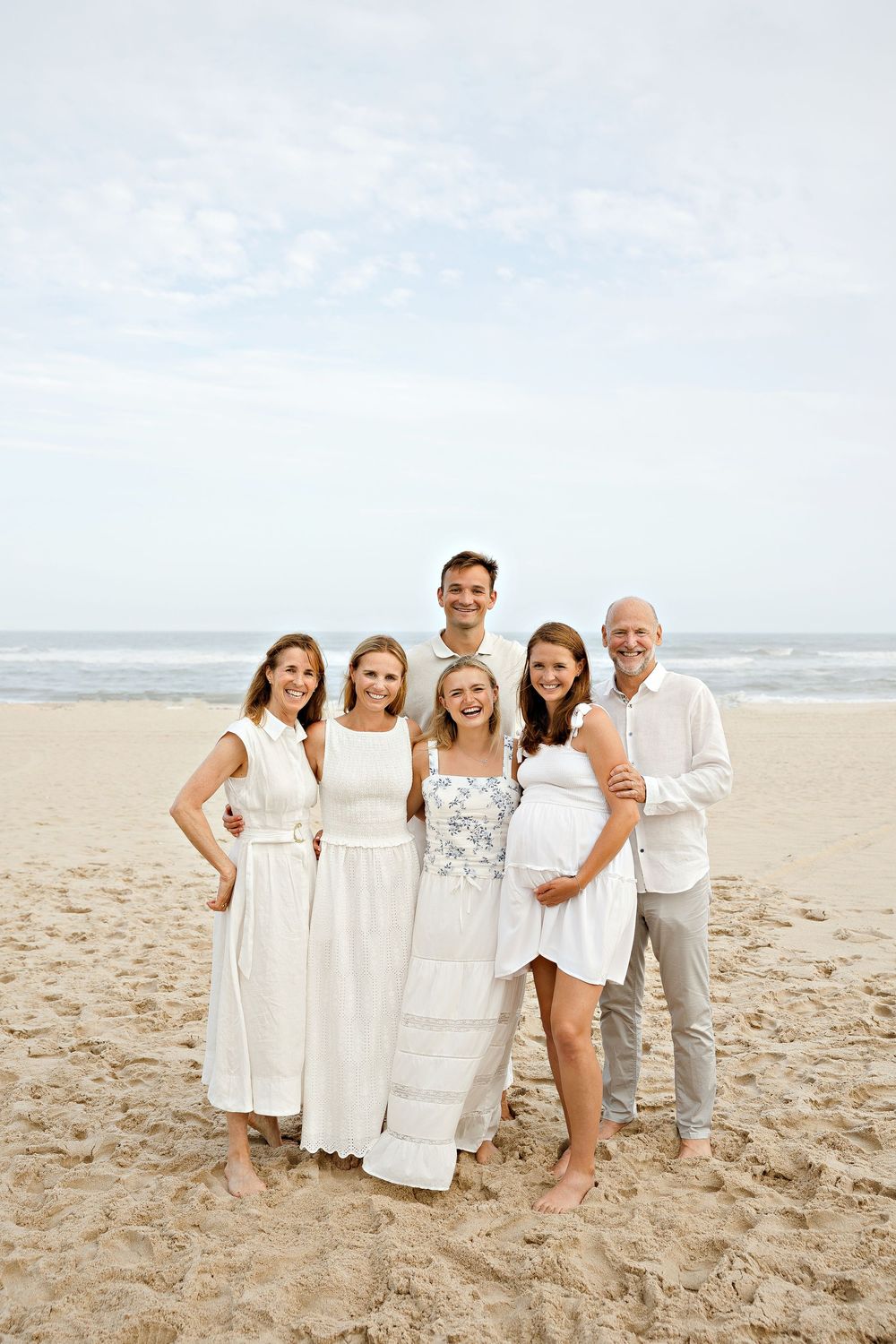 Family of six posing together on a sandy beach, wearing coordinated white outfits, with a pregnant woman smiling in front.