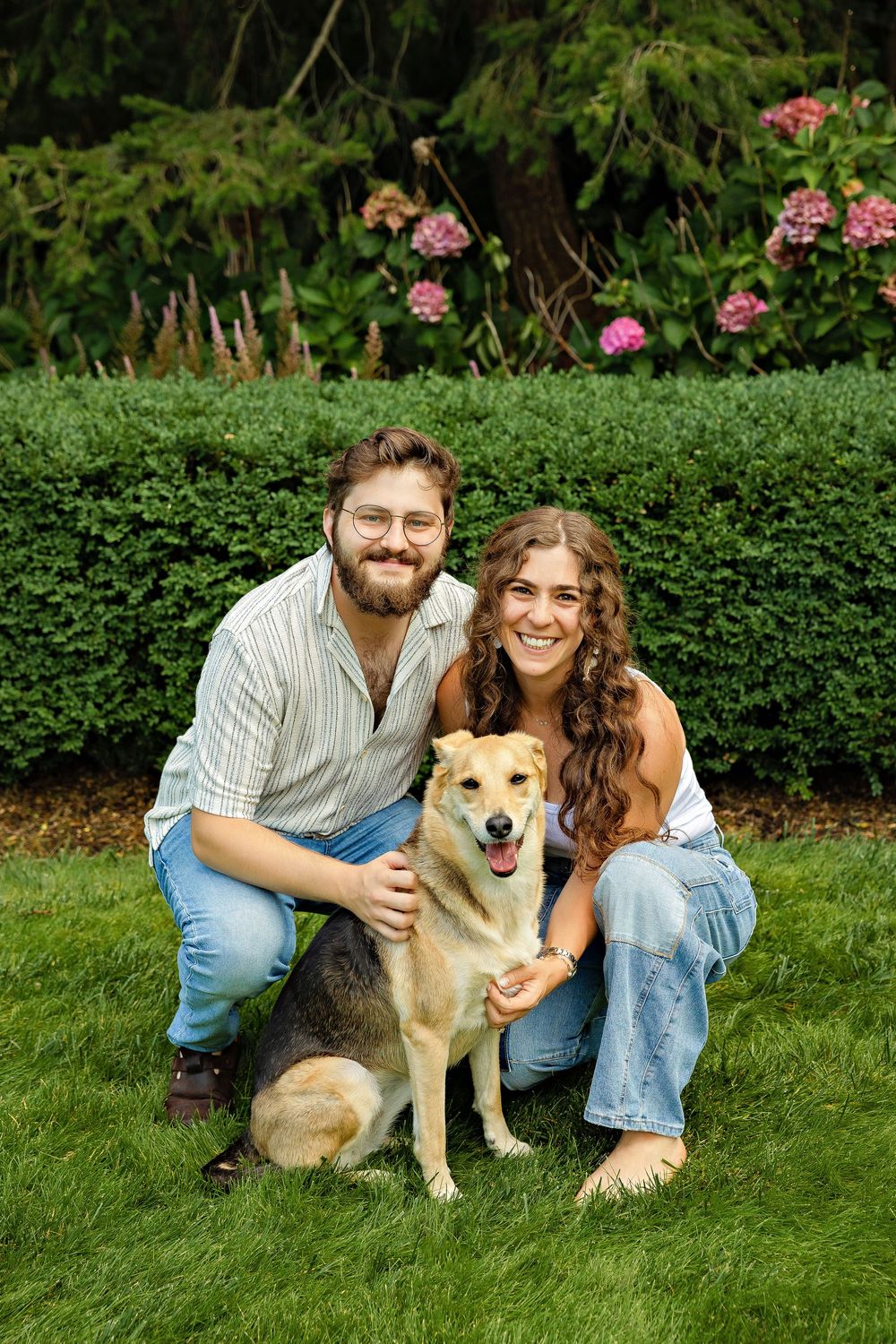 A smiling couple crouches on green grass with their happy golden dog, surrounded by lush hedges and pink flowers.