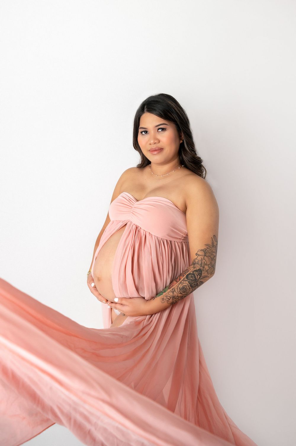 Expectant mother poses in flowing pink maternity gown against white backdrop.