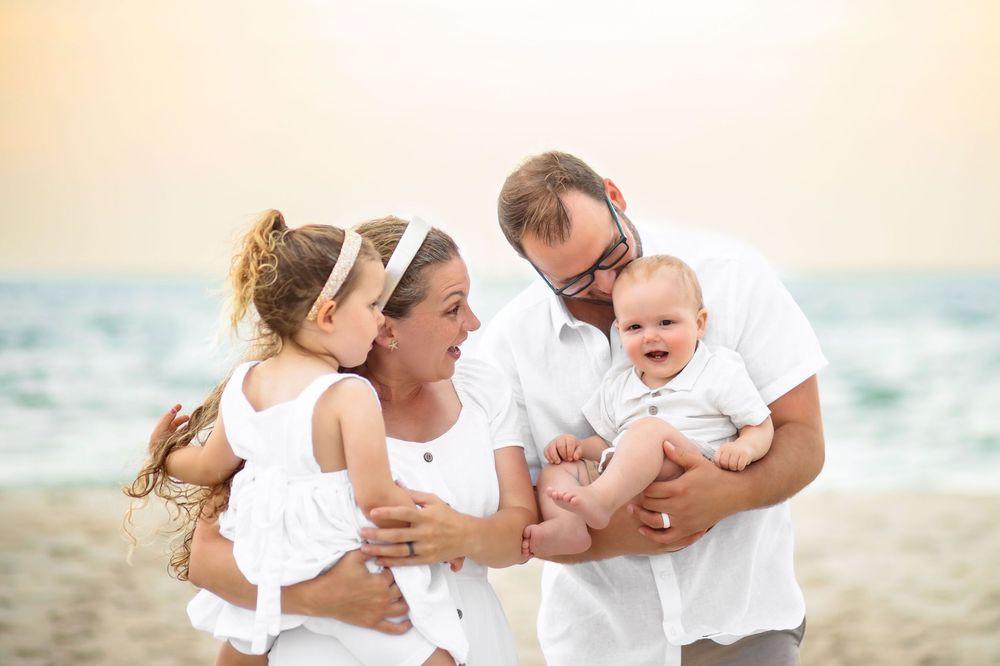 A family wearing white clothing shares joyful moments together on a sunny beach at sunset.