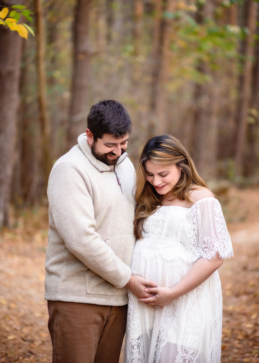 Expectant couple shares tender moment in white and beige outfits during fall forest maternity shoot.