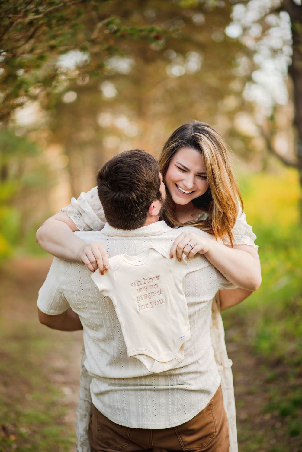 A couple embraces while holding a small onesie in a wooded setting.