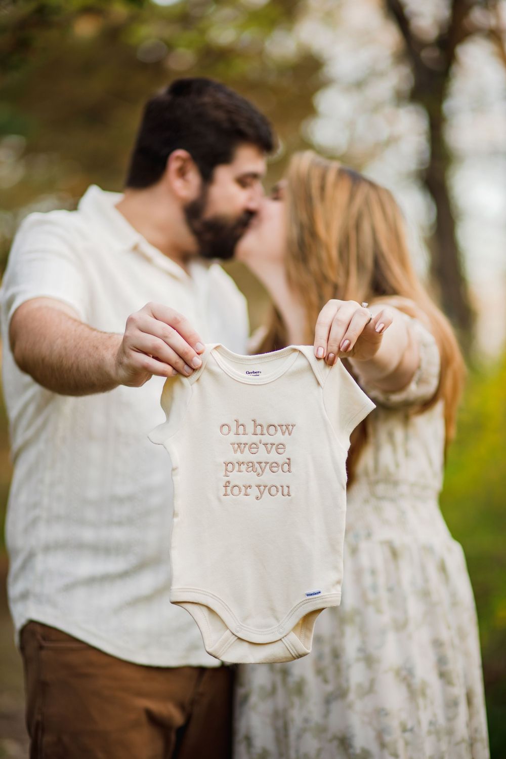 A couple shares a kiss while holding up a message-printed onesie in a forest setting.