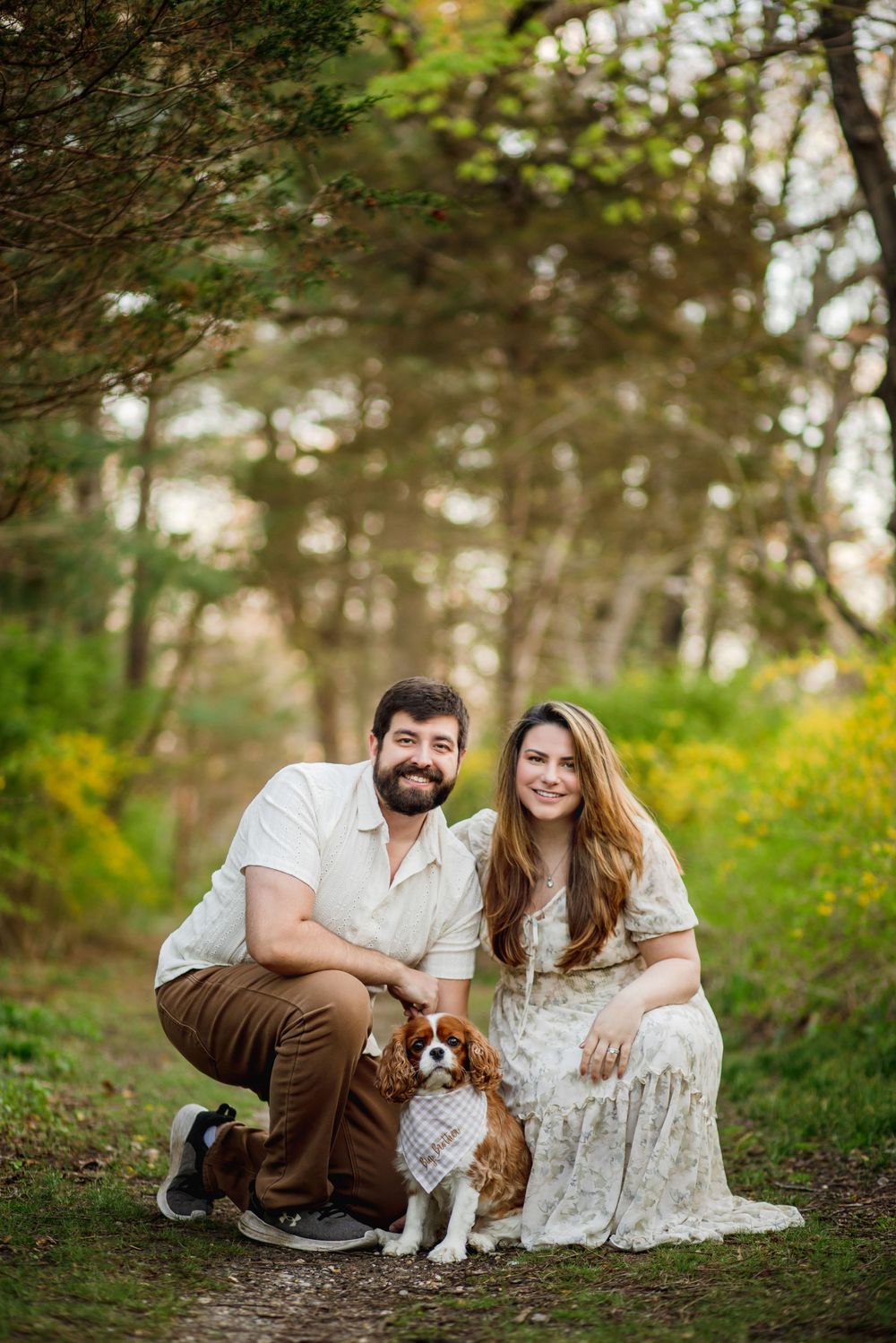 A couple in casual attire poses with their small spaniel dog on a forest trail surrounded by yellow wildflowers.