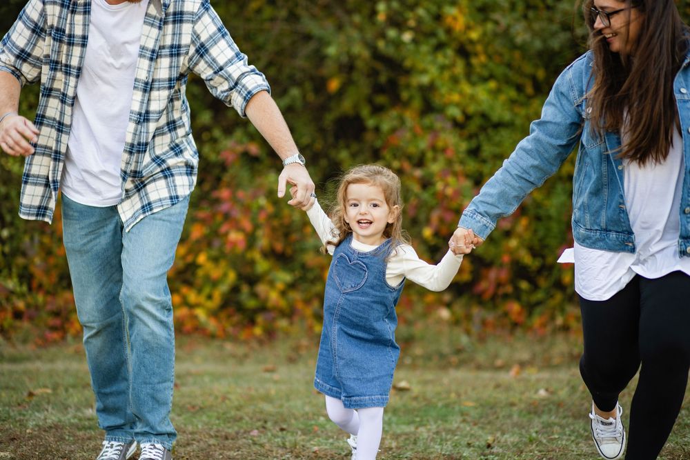A young child in denim overalls holds hands with adults while walking through a park during autumn.