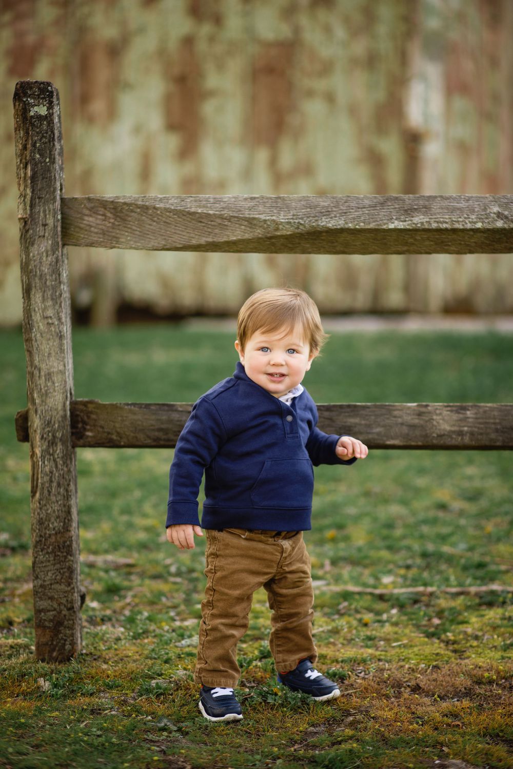Toddler in navy sweater standing by rustic wooden fence in garden setting.