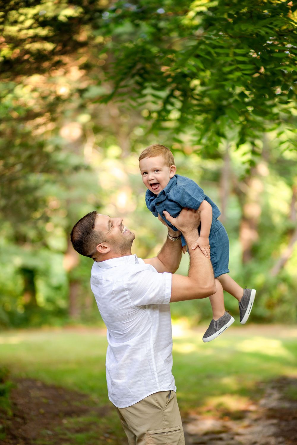 A joyful moment captured outdoors as a parent lifts a small child wearing blue into the air under green foliage.