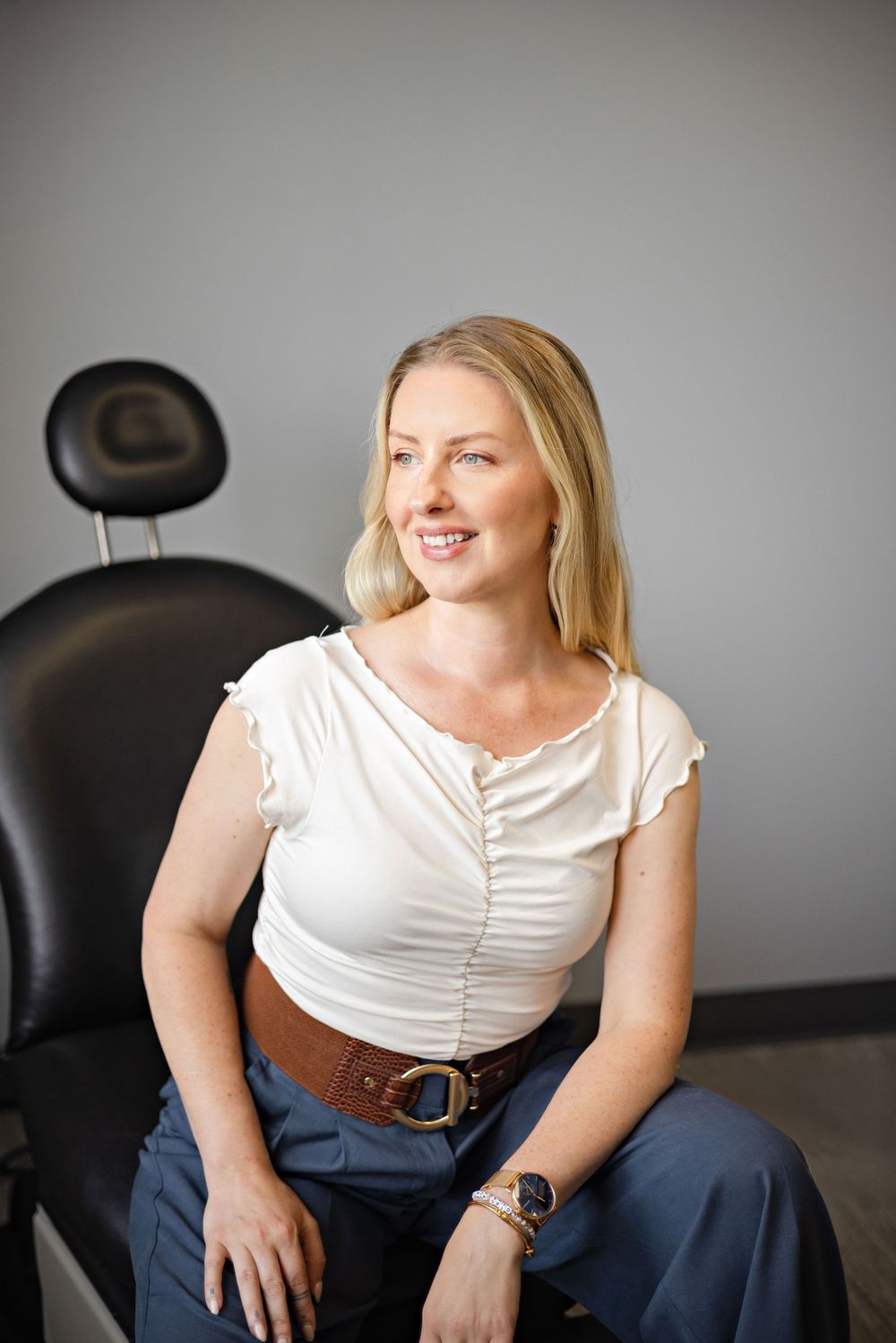 Smiling blonde woman in white top and brown belt seated in a black chair in a modern office setting.