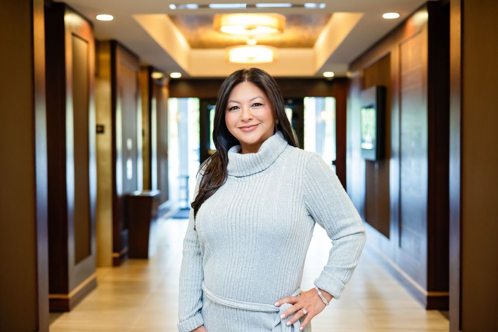 Professional in a light gray sweater stands confidently in an office hallway with warm lighting and modern decor.