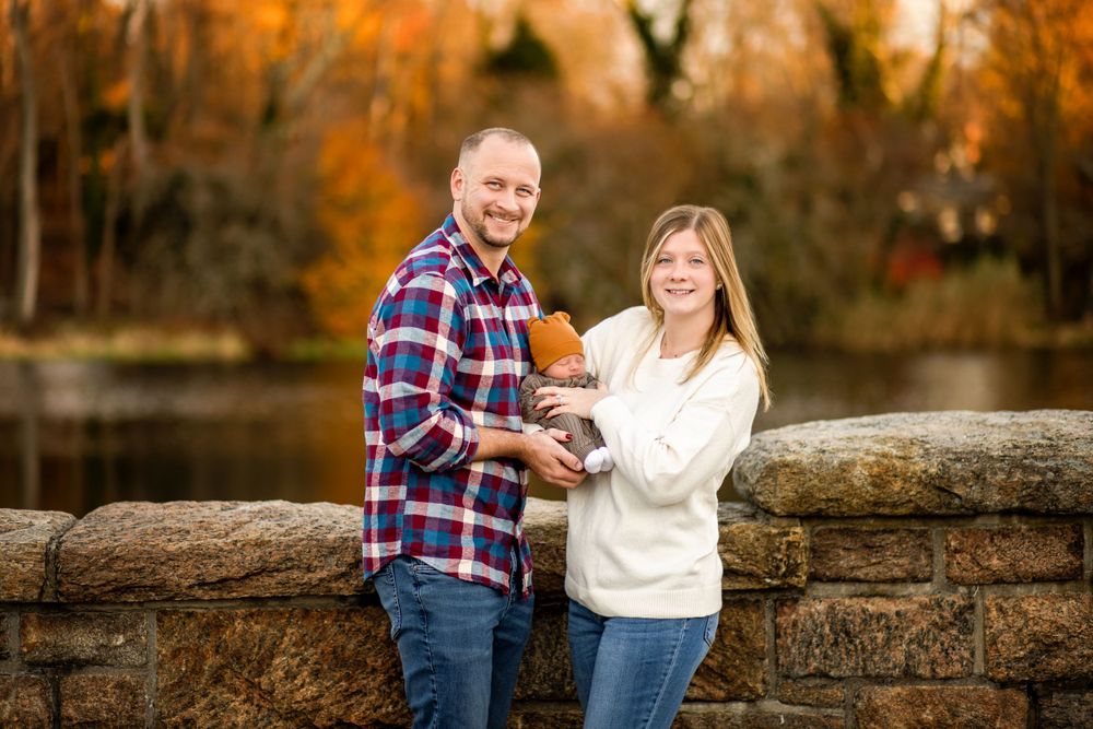 A couple stands with their baby against a stone wall during golden hour in autumn.