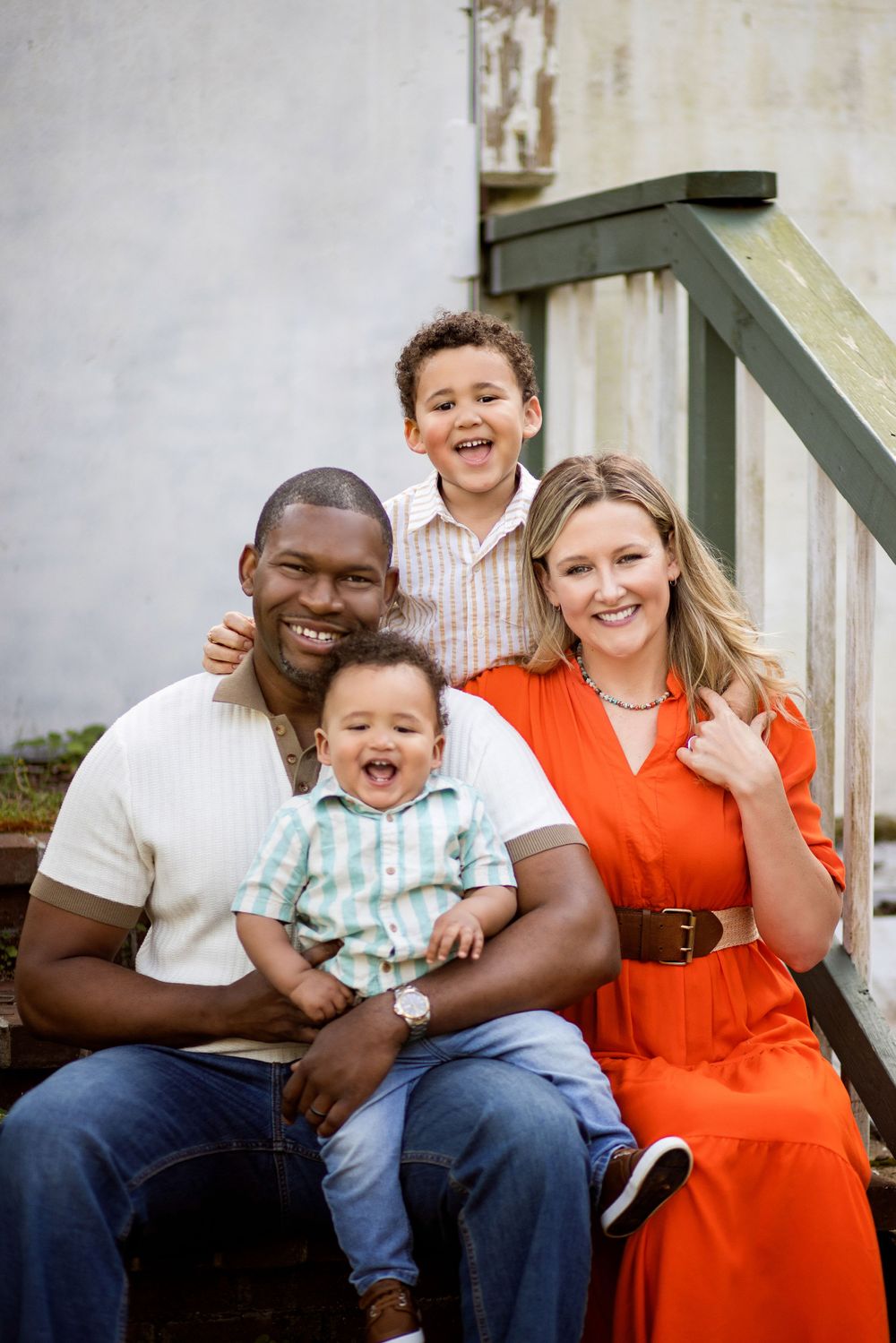 A happy family of four sits together on outdoor steps, with bright smiles and casual attire.