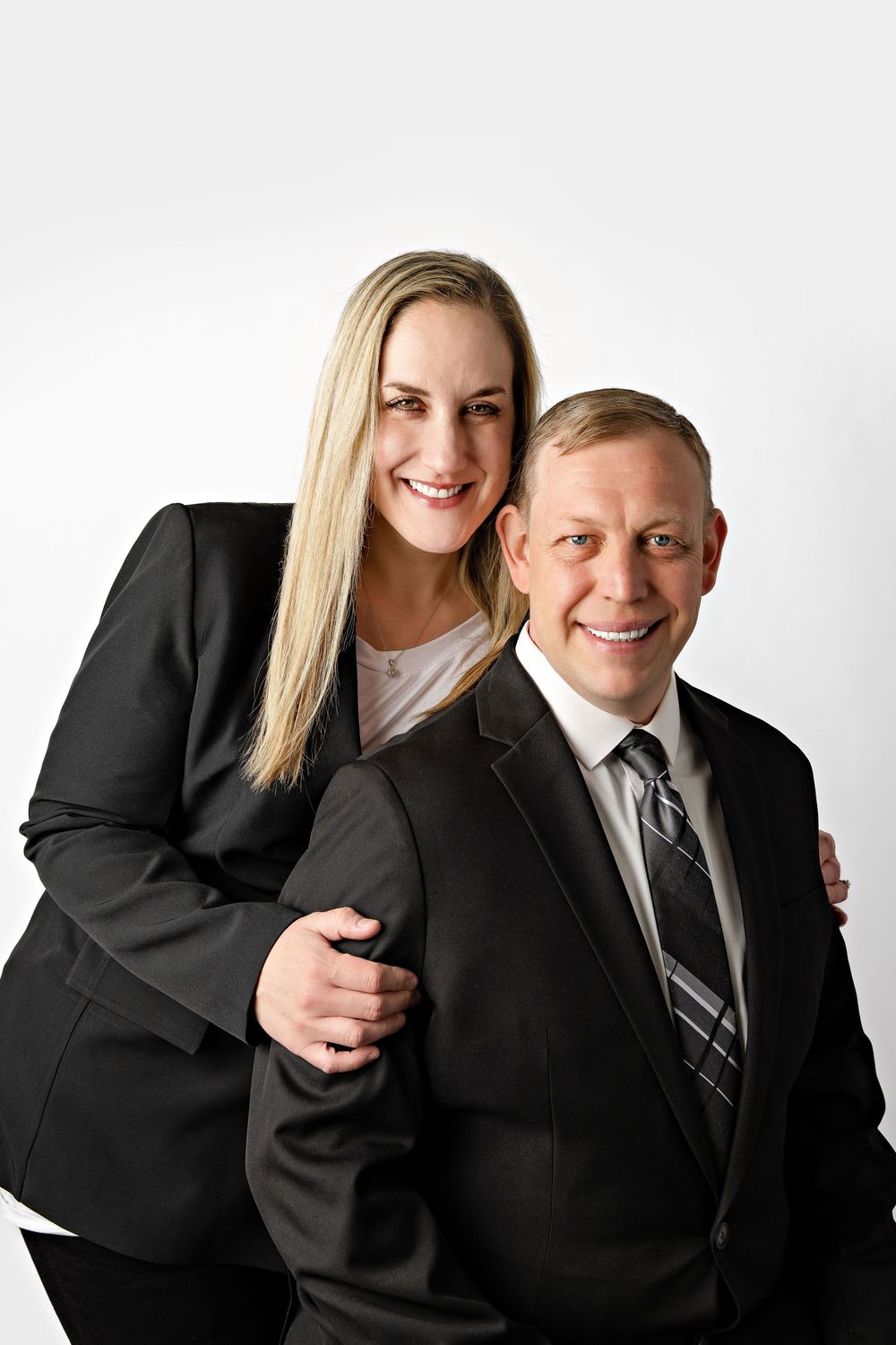 Professional couple in dark suits smiling together against a white background in a formal portrait photo.