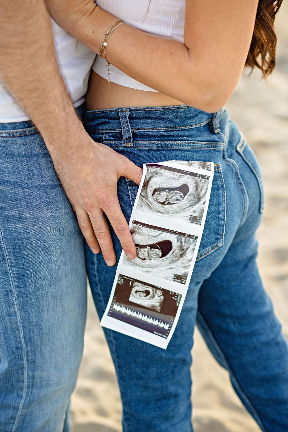 Couple holding ultrasound sonogram photos in jeans pocket on sandy beach, announcing pregnancy.