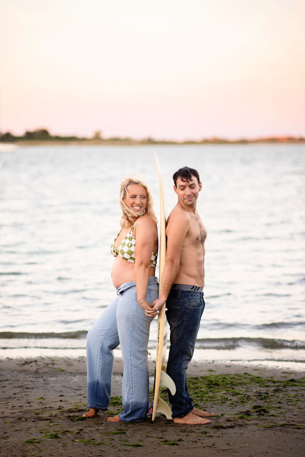 Couple posing together on beach with surfboard at sunset.