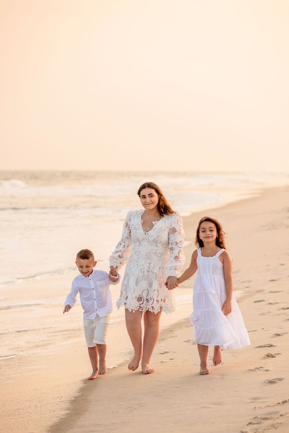 Family walking hand in hand along beach shoreline at sunset in white outfits.