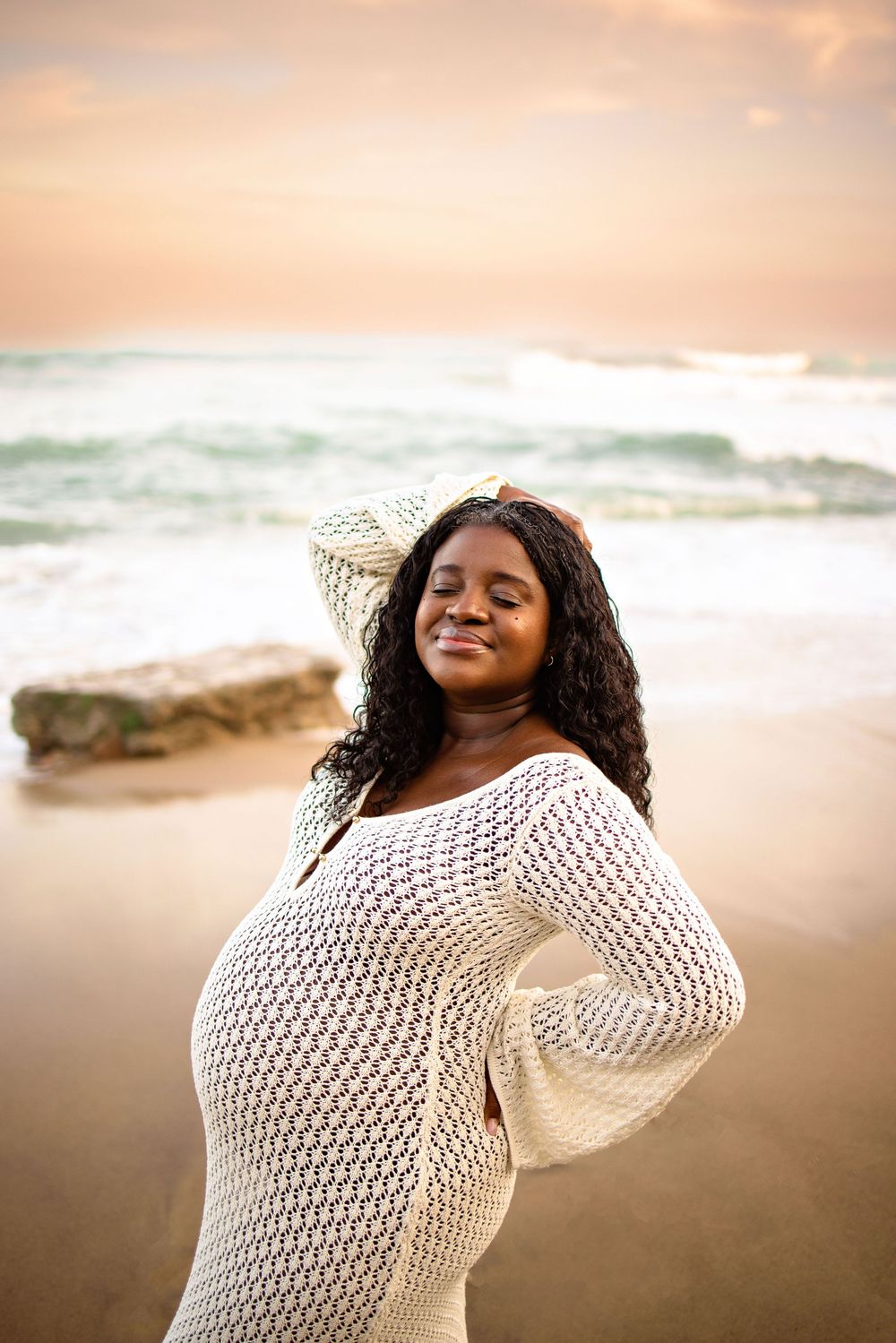 A person in a white crochet dress poses confidently on a beach at sunset with waves crashing in the background.
