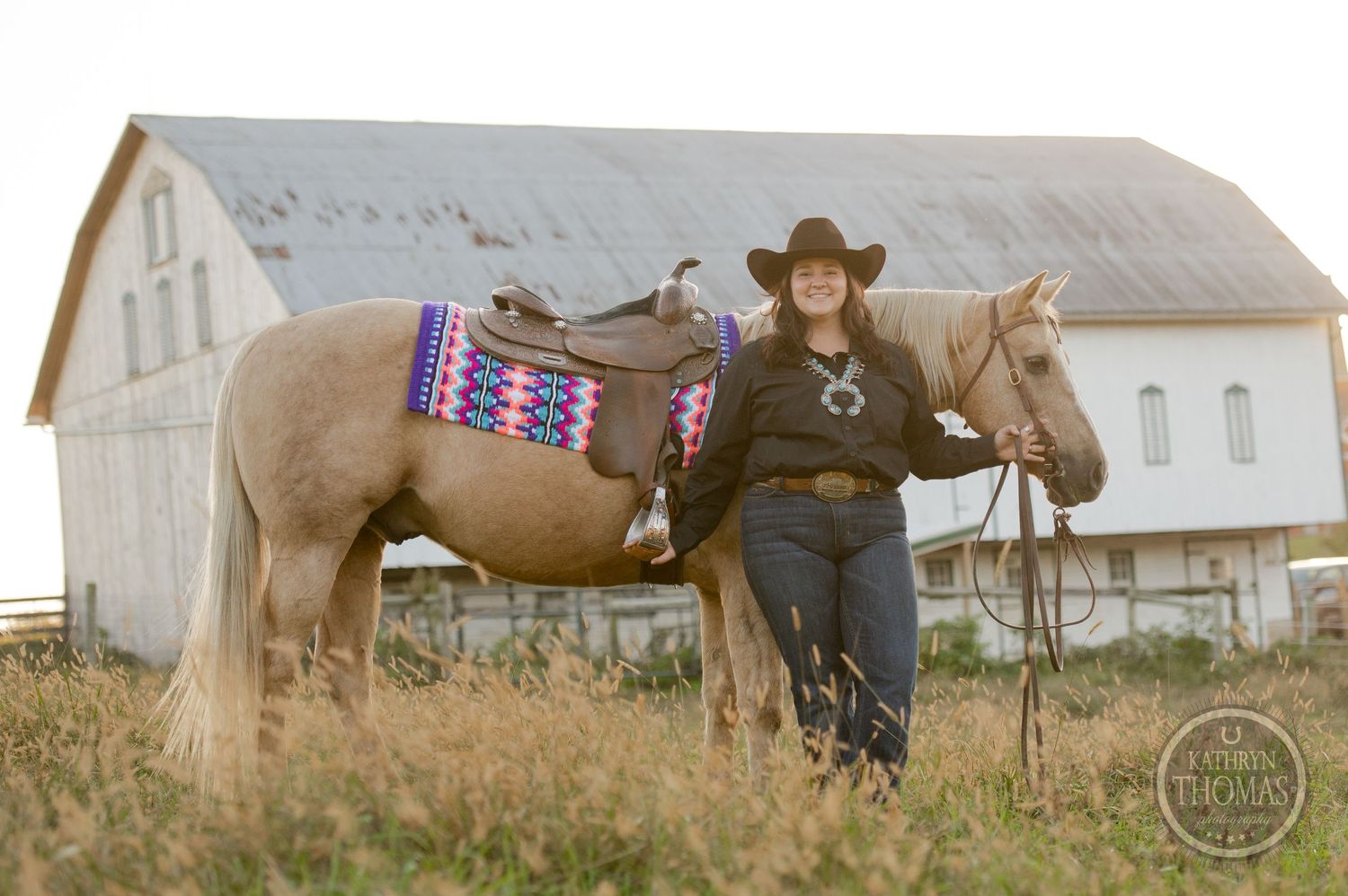 Pale horse with decorative western saddle pad stands near white barn building.