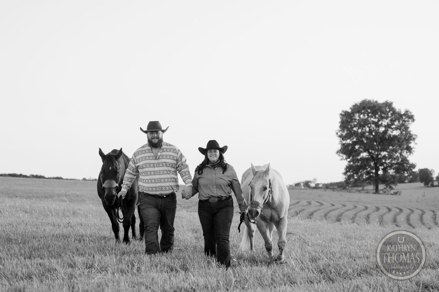 Black and white image of two people walking with horses through a rural field.