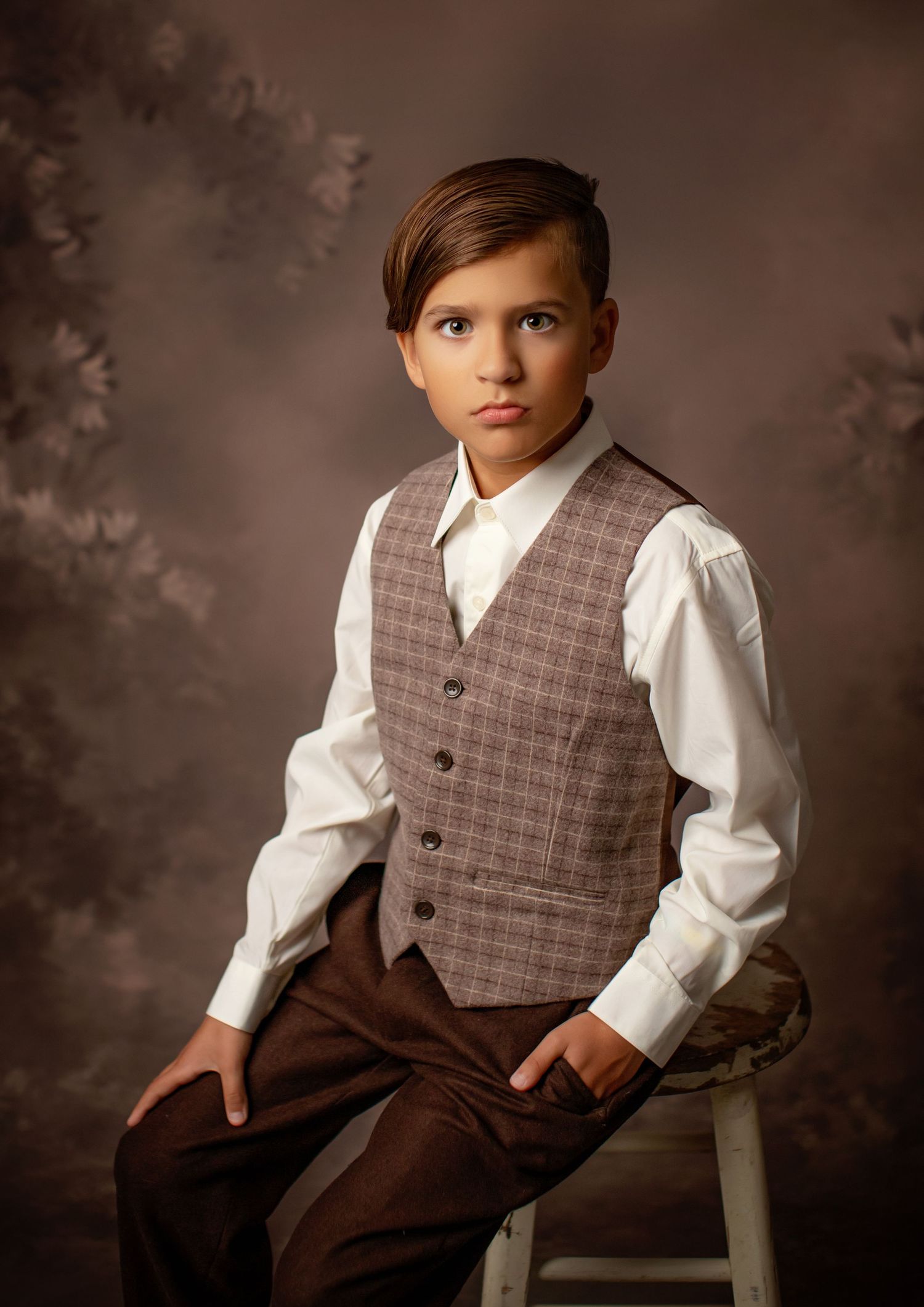Studio portrait of a child in classic brown attire with plaid vest and white dress shirt sitting on wooden stool.