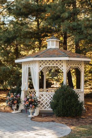 Elegant white lattice gazebo decorated with colorful floral arrangements surrounded by tall pine trees at sunset.
