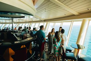 Guests socializing at a cruise ship bar lounge with panoramic ocean views through large windows.