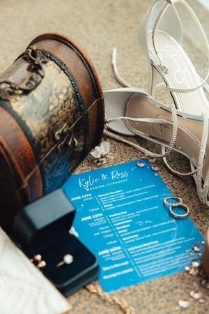 Wedding flat lay featuring blue itinerary cards, bridal heels, rings, and vintage purse on textured surface.