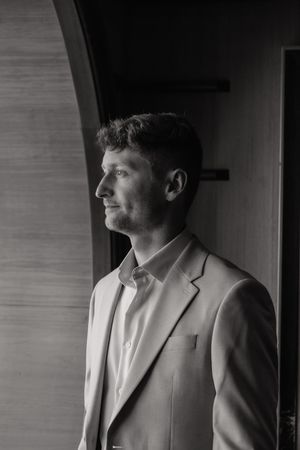 Elegant black and white portrait of groom in suit gazing thoughtfully out window in soft light.